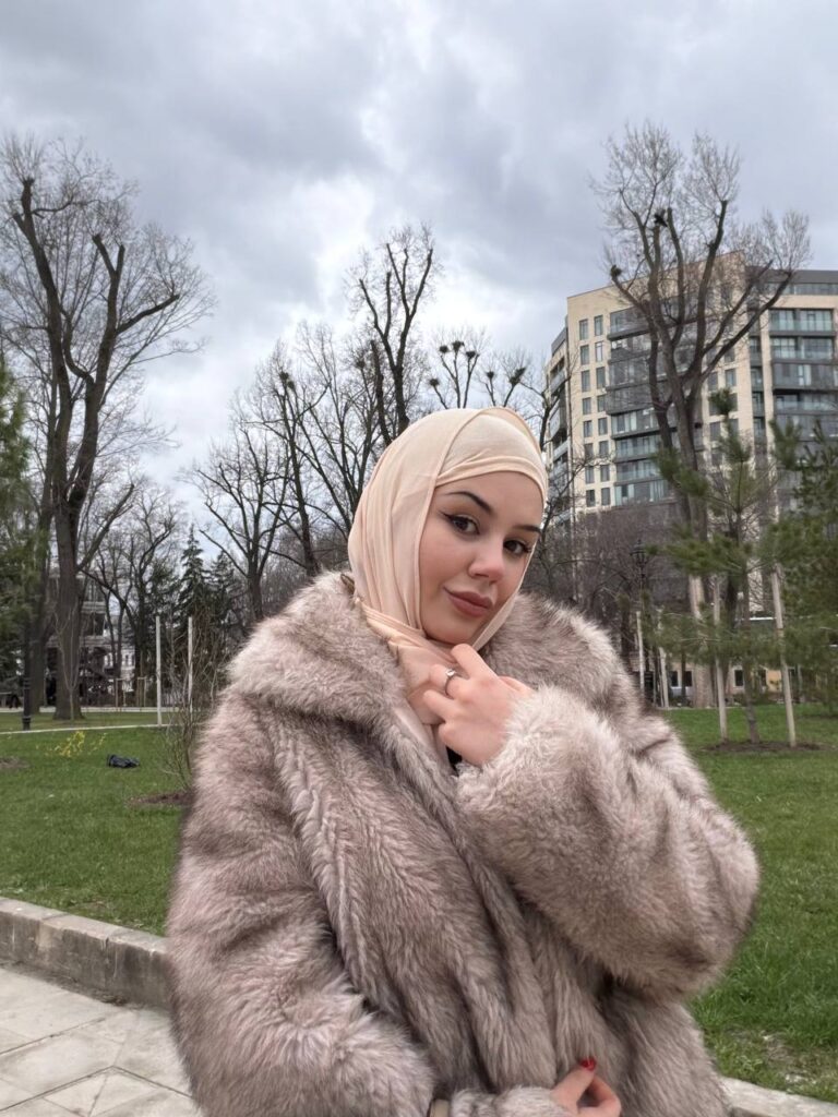 Woman in a beige hijab and fluffy fur coat posing in a park with bare trees and modern buildings behind her.