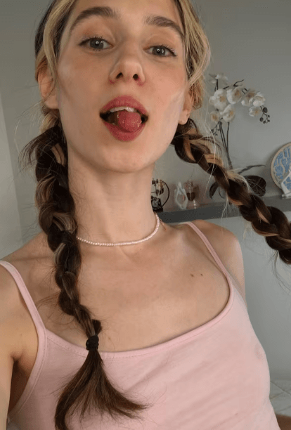 Young woman with two braids and a pink tank top sticking out her tongue in a playful pose, background shows a shelf and flowers.