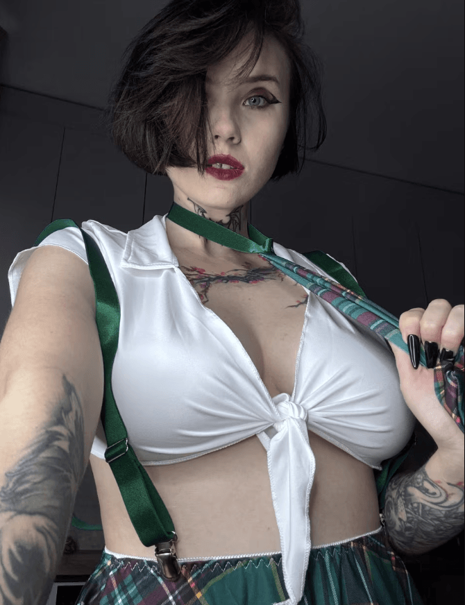 Self-portrait of a tattooed woman with dark hair, wearing a knot-front white top, green suspenders, and a plaid skirt, facing the camera.