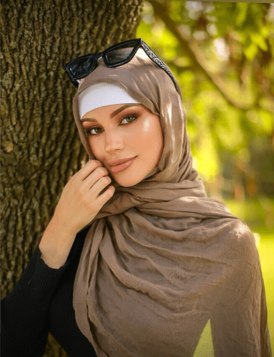 Portrait of a woman wearing a beige hijab and white headband, sunglasses resting on her head, leaning against a tree in a sunny park.