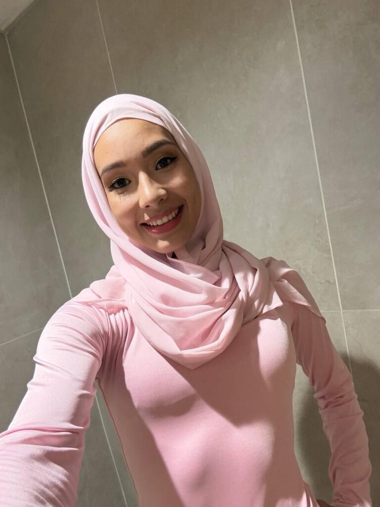 Young woman wearing a light pink hijab and matching long-sleeve top taking a selfie and smiling at the camera against a tiled wall backdrop.