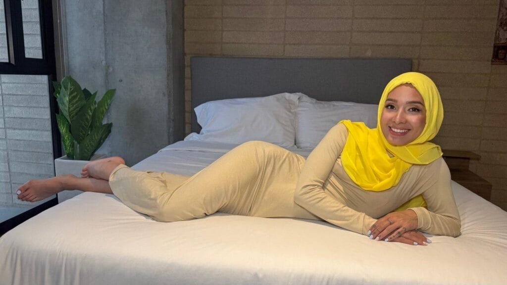Smiling woman in a beige outfit and bright yellow hijab lying on a white bed in a modern bedroom.