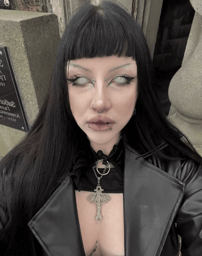 Person with black hair and bangs wearing a black leather jacket, a large ornate cross pendant, and multiple facial piercings with pale makeup.