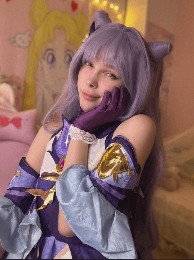 Cosplayer in a purple and blue fantasy costume with lavender wig and horn headband, posing with hand on the cheek in a pastel room.