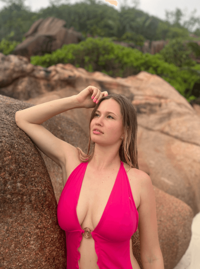 Woman in a bright pink halter swimsuit leaning against large brown rocks, with green shrubbery in the background on a sunny day.