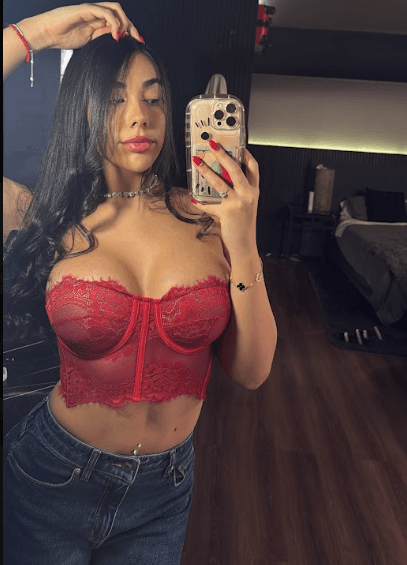 Young woman taking a mirror selfie in a red lace bustier and jeans in a bedroom/dressing area, phone visible in hand and long dark hair down.
