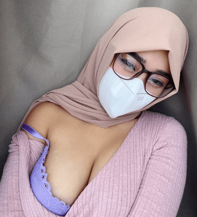 Selfie of a woman wearing a beige hijab and mask with glasses, posing in a pink sweater and a purple lace bra visible at the neckline.
