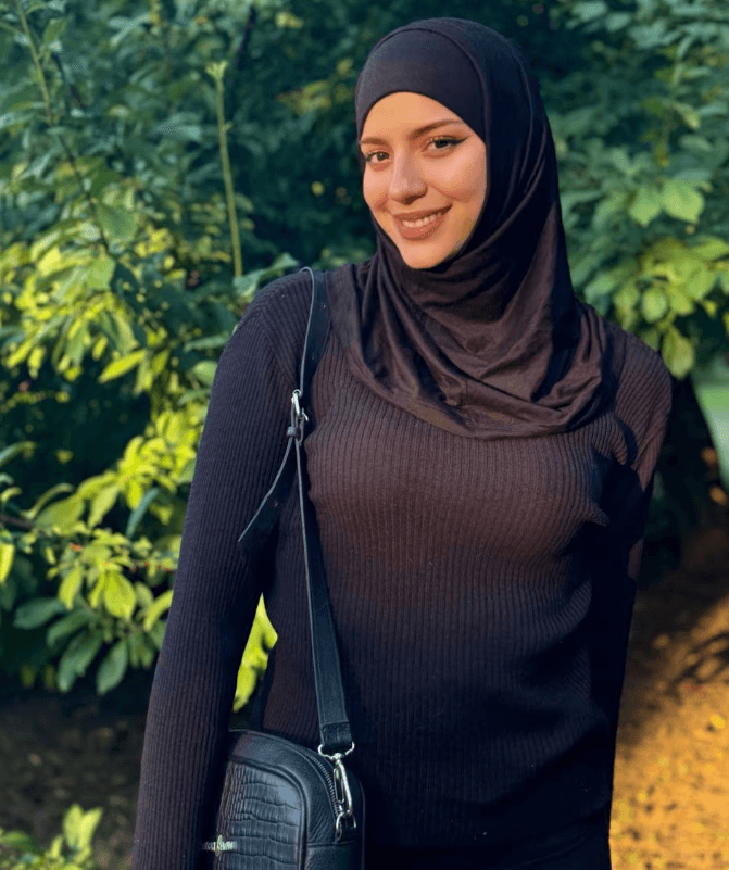 Woman wearing a black hijab and ribbed sweater stands outdoors with greenery in the background, carrying a black crossbody bag.
