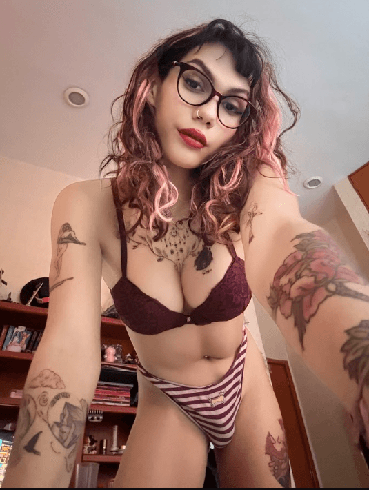Self-portrait of a person in burgundy lace lingerie and striped bottoms, with pink-tinted hair, glasses, red lipstick, and visible arm/chest tattoos.