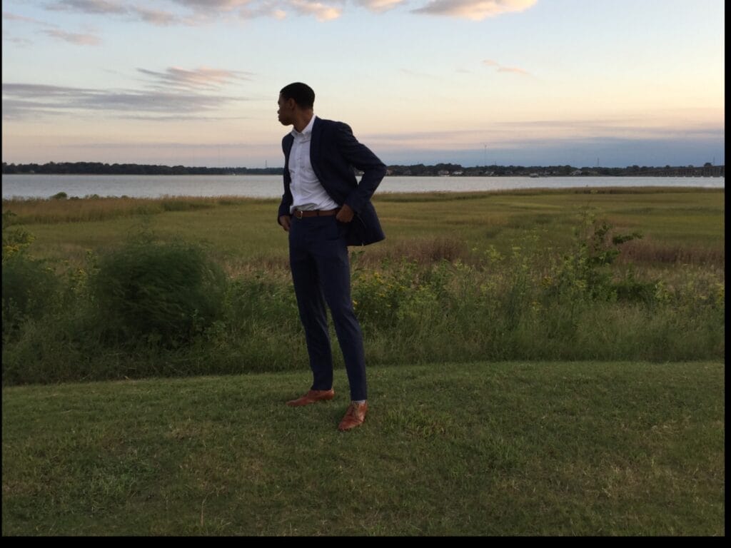 Man in a navy suit standing in a grassy field beside a lake at sunset, looking to the left.