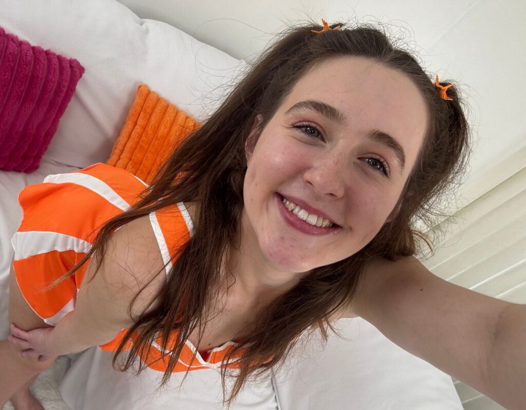Smiling woman with orange hair clips lying on a white bed, wearing an orange and white striped top.