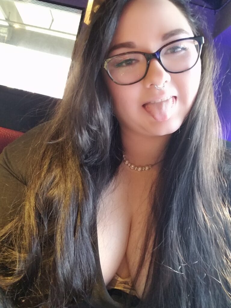 Close-up portrait of a young woman with long dark hair and glasses making a playful face by sticking out her tongue, wearing a beaded necklace.