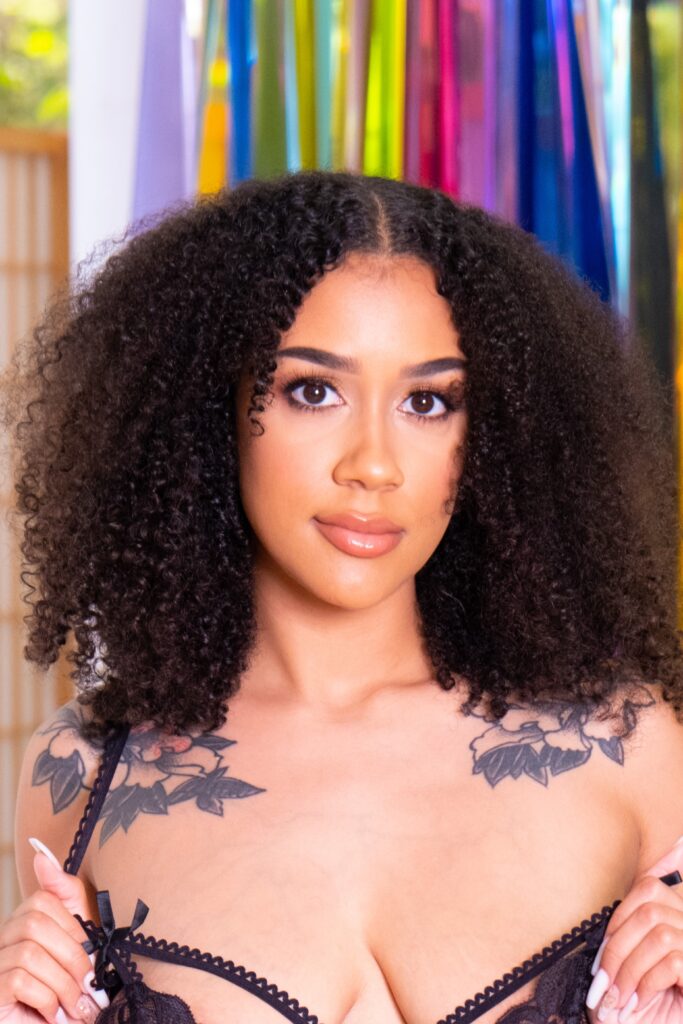 Close-up portrait of a woman with voluminous curly hair, wearing black lace lingerie, with colorful ribbons in the background and visible shoulder tattoos.