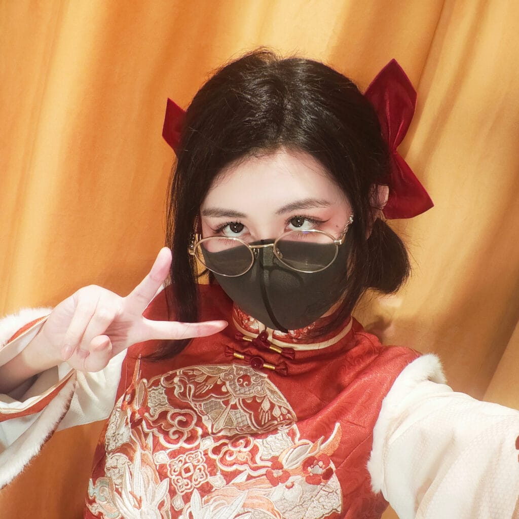 Woman in a red and cream embroidered kimono with a red hair bow, making a peace sign beside glasses against an orange curtain.