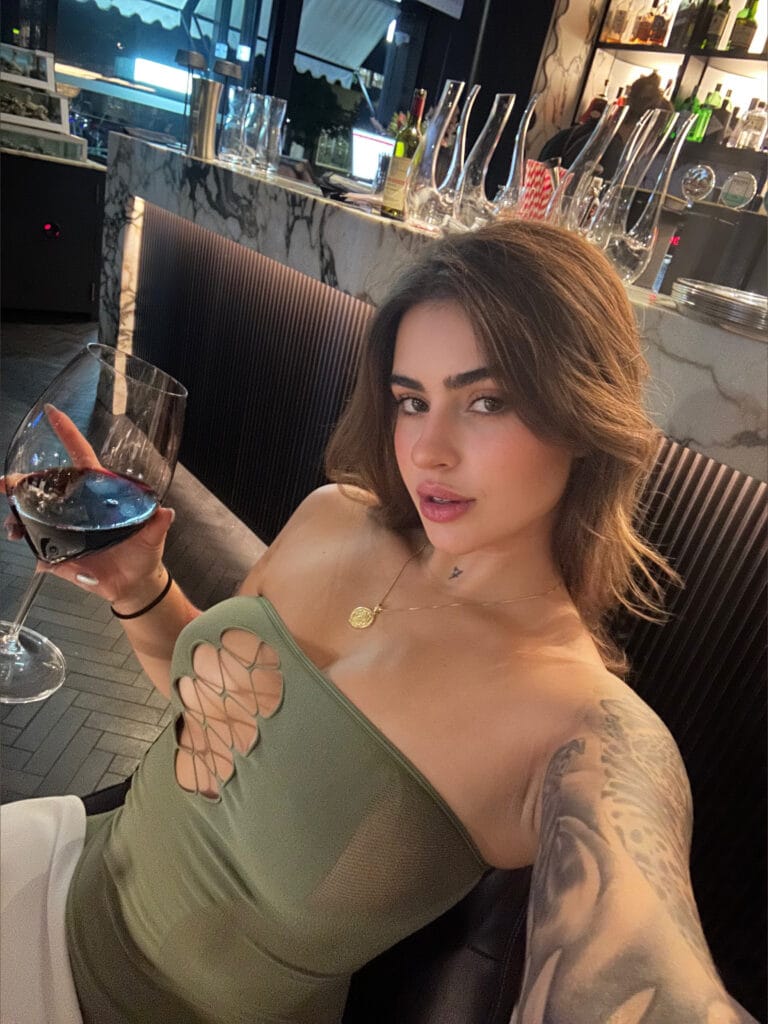 Selfie of a woman in a green cut-out dress at a marble-bar lounge, holding a glass of red wine.