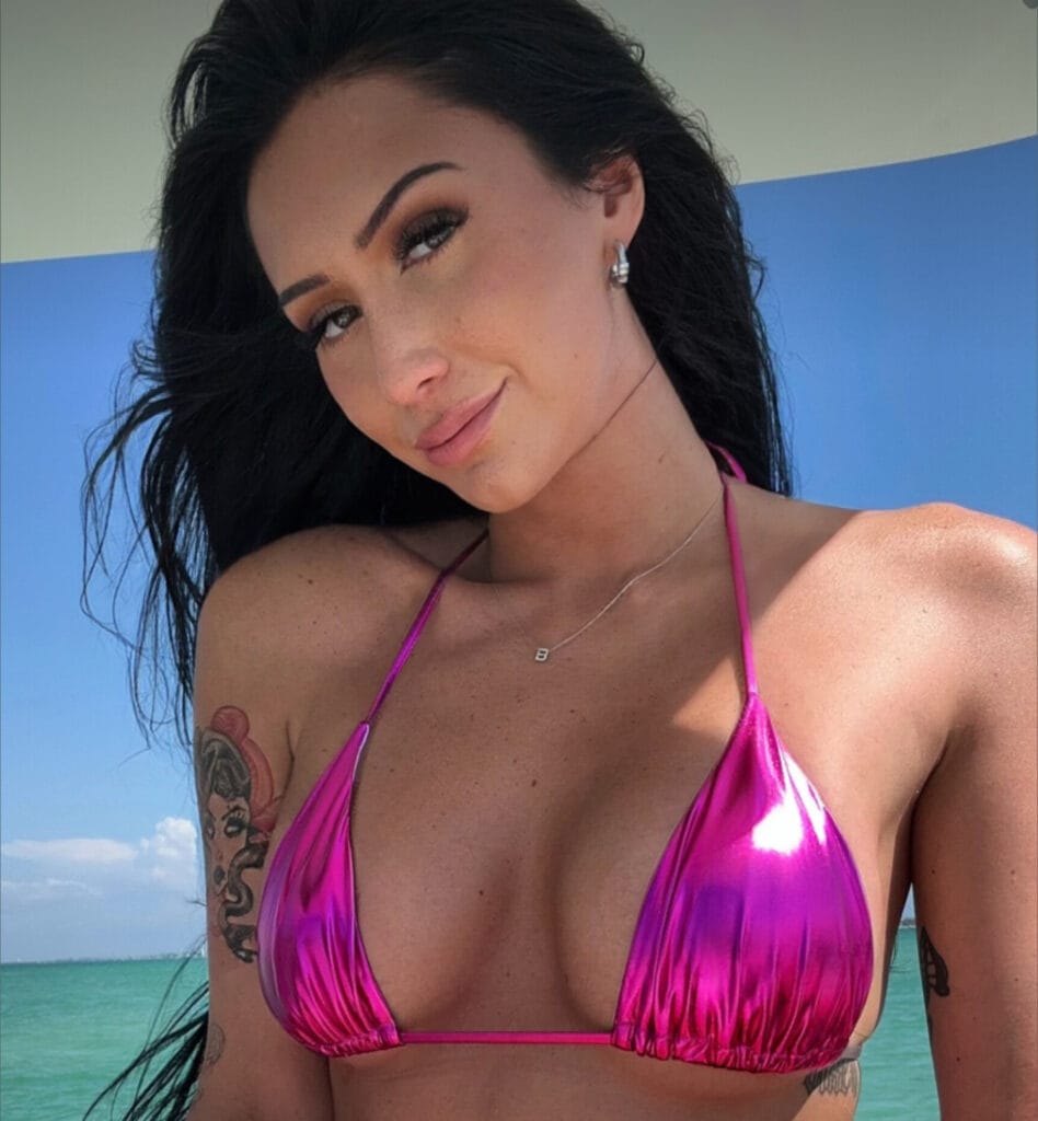 Close-up of a woman in a pink bikini at the beach, turquoise water behind her, with a necklace and visible tattoos on her arm.