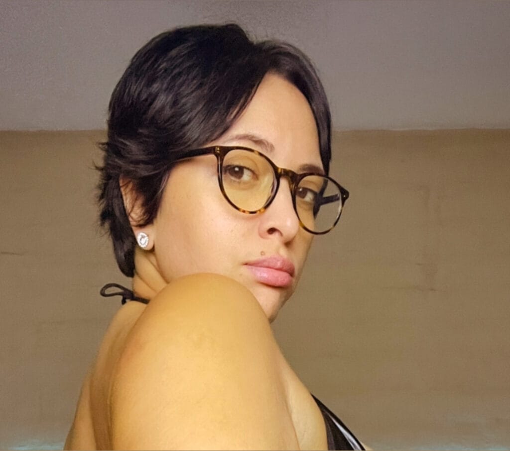 Close-up portrait of a person with short dark hair, tortoiseshell glasses, and diamond stud earrings facing left.