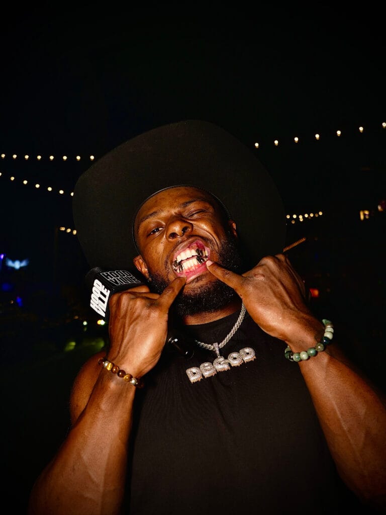 Man wearing a wide-brimmed black hat and grillz smiles at the camera during a night event, holding a microphone near his mouth with string lights in the background.