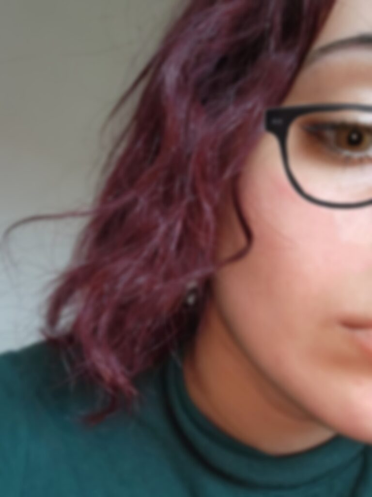 Close-up of a person wearing black glasses and purple hair, showing part of the face and a teal shirt collar