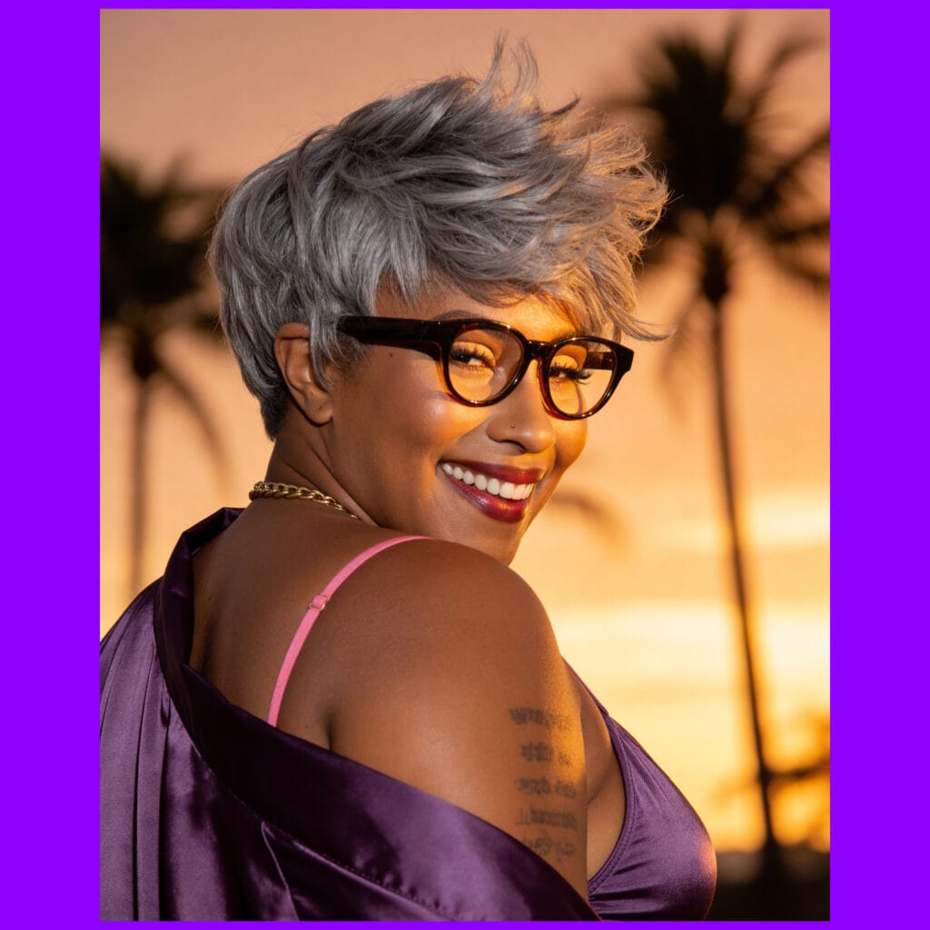 Person with short gray hair and dark glasses smiles at sunset, wearing a purple satin dress; palm trees in the background; tattoos visible on the chest area.