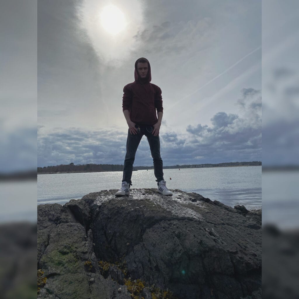 Man in a burgundy hoodie stands on jagged rocks at the water's edge under a cloudy sky with the sun behind him.