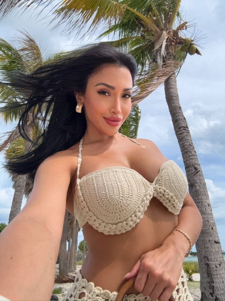 Selfie of a woman in a beige crochet bikini top on a sunny beach with palm trees in the background.