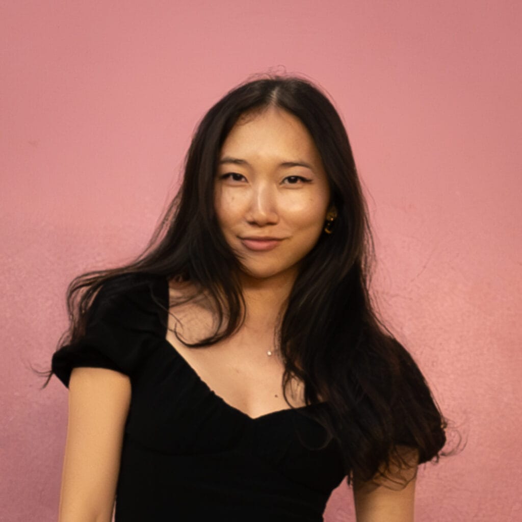 Portrait of a woman with long dark hair wearing a black top, standing against a pink background.