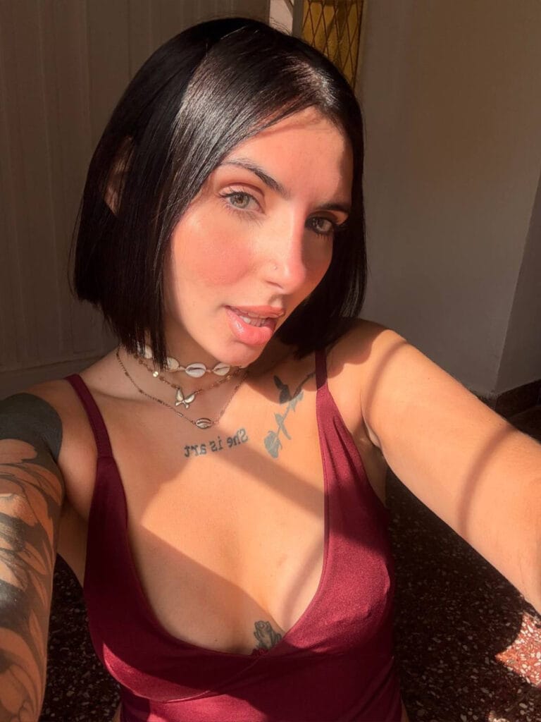 Self-portrait of a woman with short black hair wearing a burgundy dress, accessorized with shells necklaces, with tattoos visible on her chest and arms, bathed in warm sunlight.