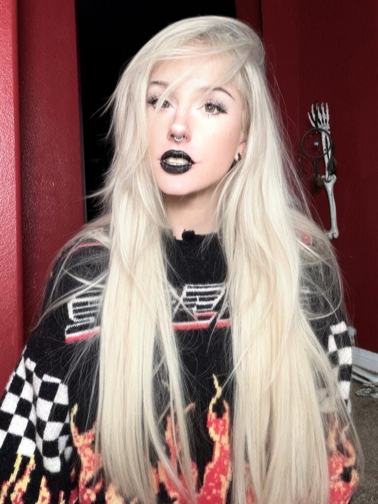 Portrait of a woman with long platinum blonde hair, black lipstick, nose ring, wearing a black-and-white patterned sweater, against a red wall.