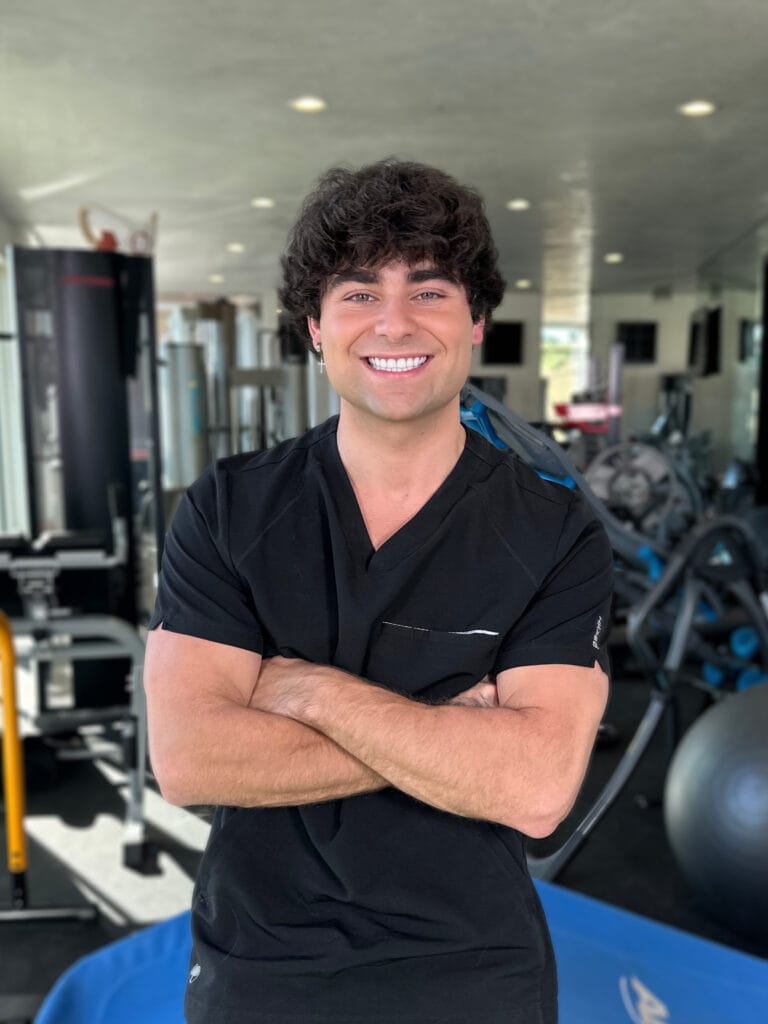Smiling man in black scrubs with arms crossed in a gym, fitness equipment visible in the background