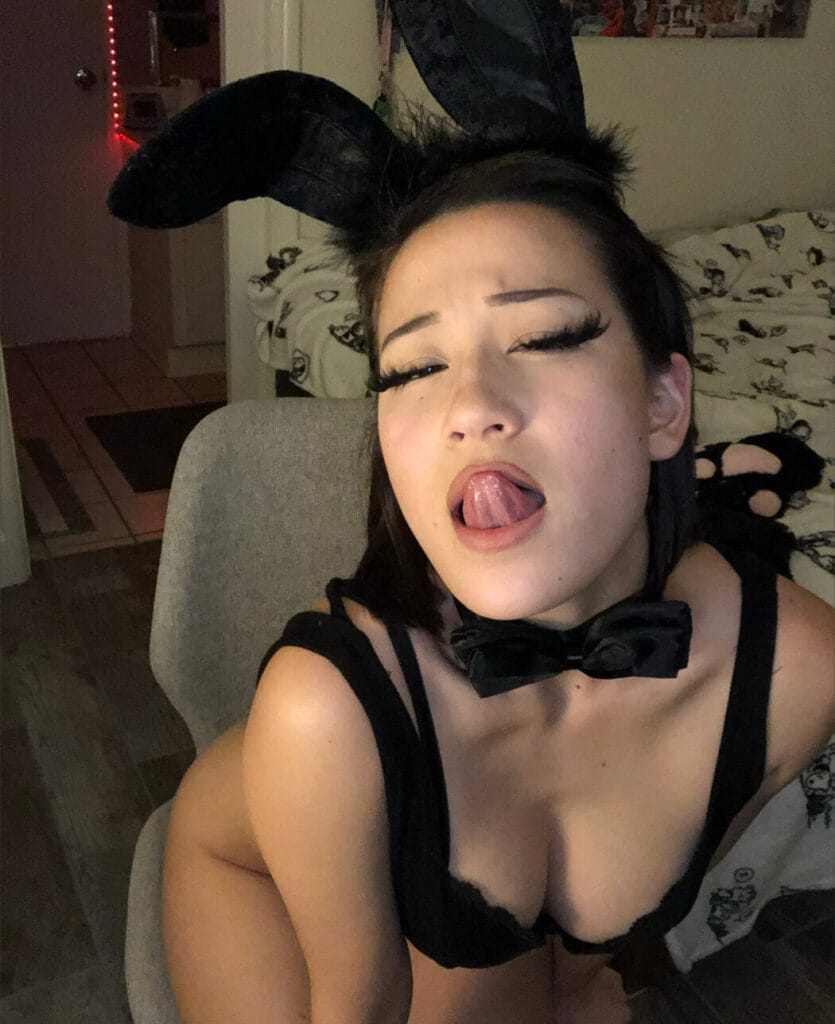 Woman in a black bunny costume with tall ears and bow tie, posing playfully with her tongue out at the camera in a dimly lit room.