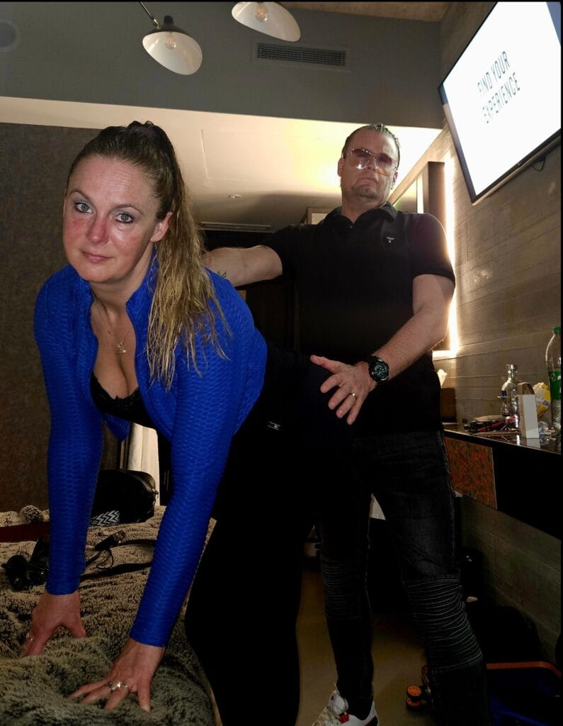 Woman in a bright blue jacket leaning on a furry surface with a man in a black polo standing behind her indoors.