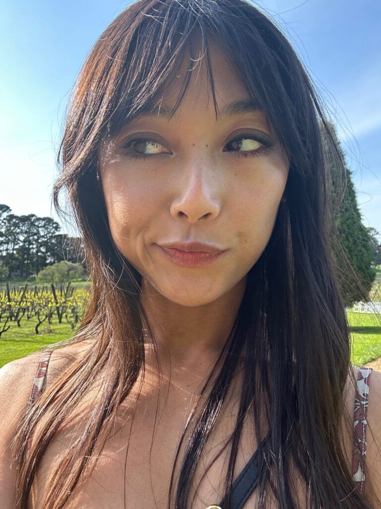 Close-up portrait of a woman with long dark hair outdoors on a sunny day, looking to the side with a subtle smile, wearing a necklace.